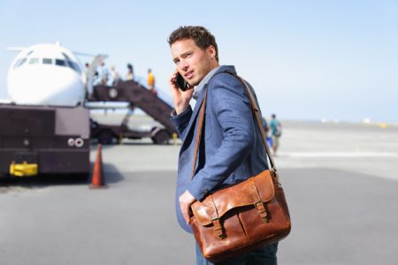 Airport business man on smartphone by plane. Young male professional hip businessman talking on smartphone boarding airplane going flying on business trip. Casual male wearing suit and laptop bag.